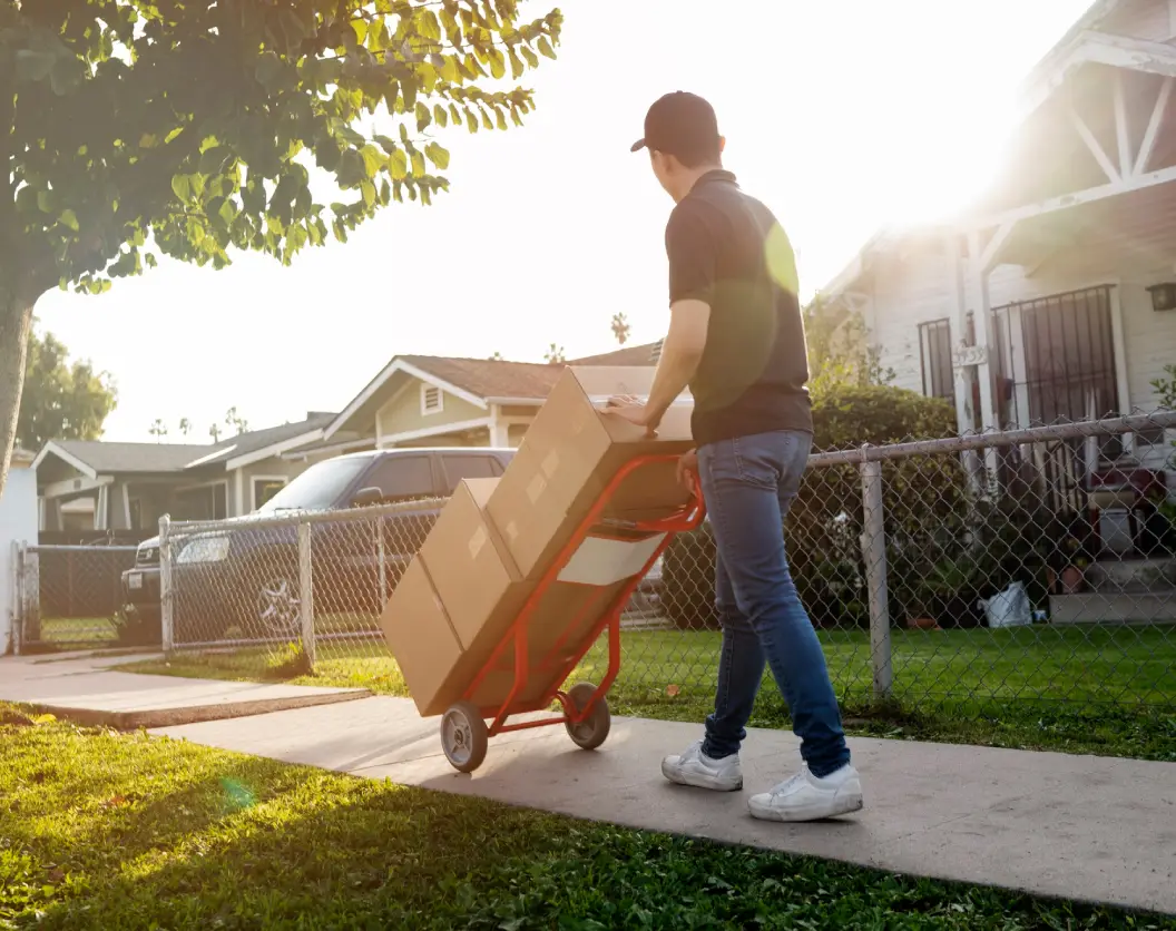 A delivery person pushes a red dolly loaded with boxes along a sidewalk, with houses and sunlight in the background