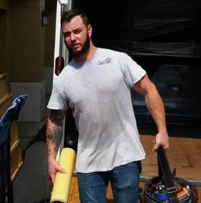 A person carries a rolled yellow tube and a bag of tools while standing in a delivery truck, with a cart nearby