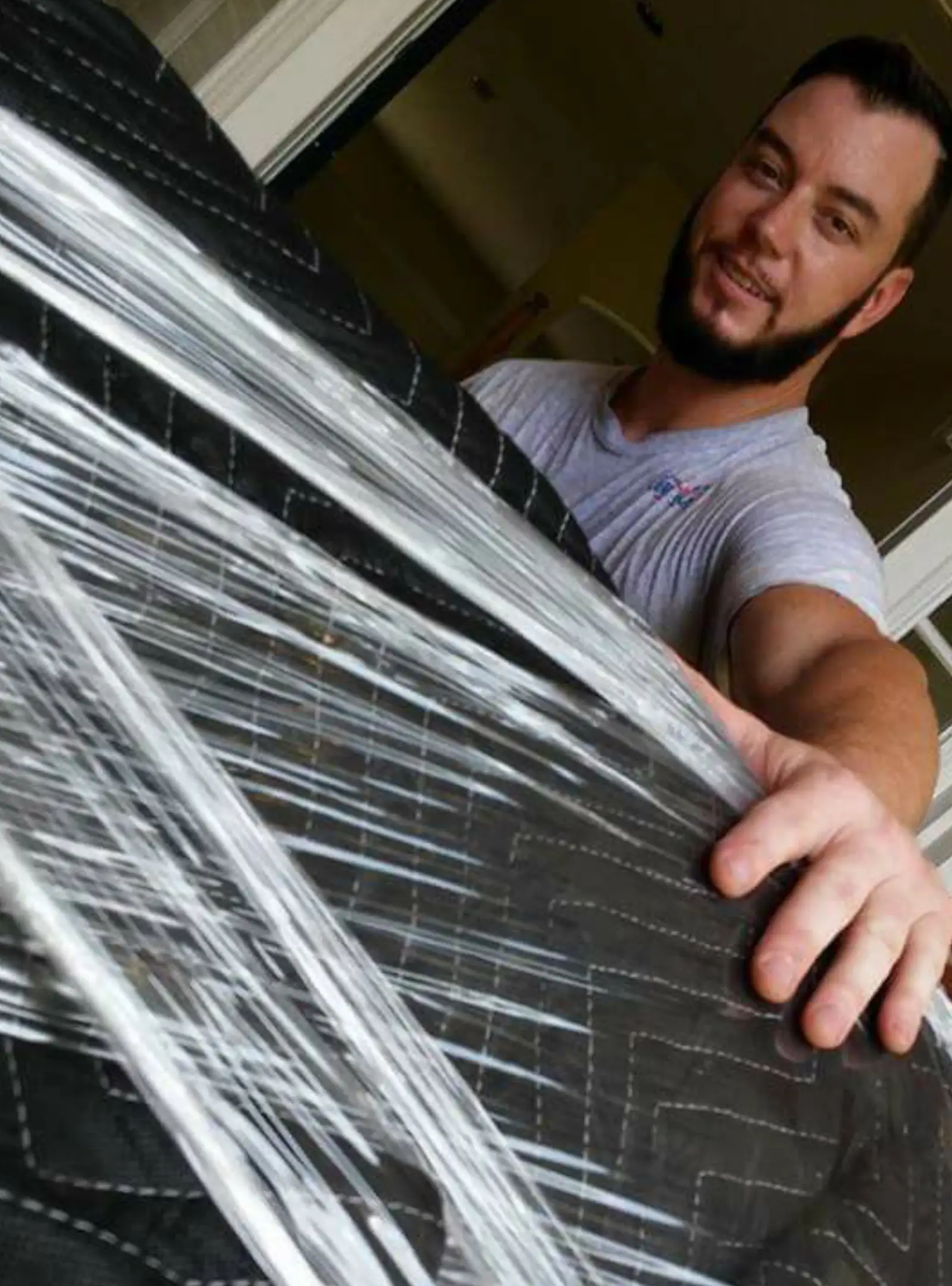 A person is securing a large, black padded object with clear plastic wrap, preparing it for transport